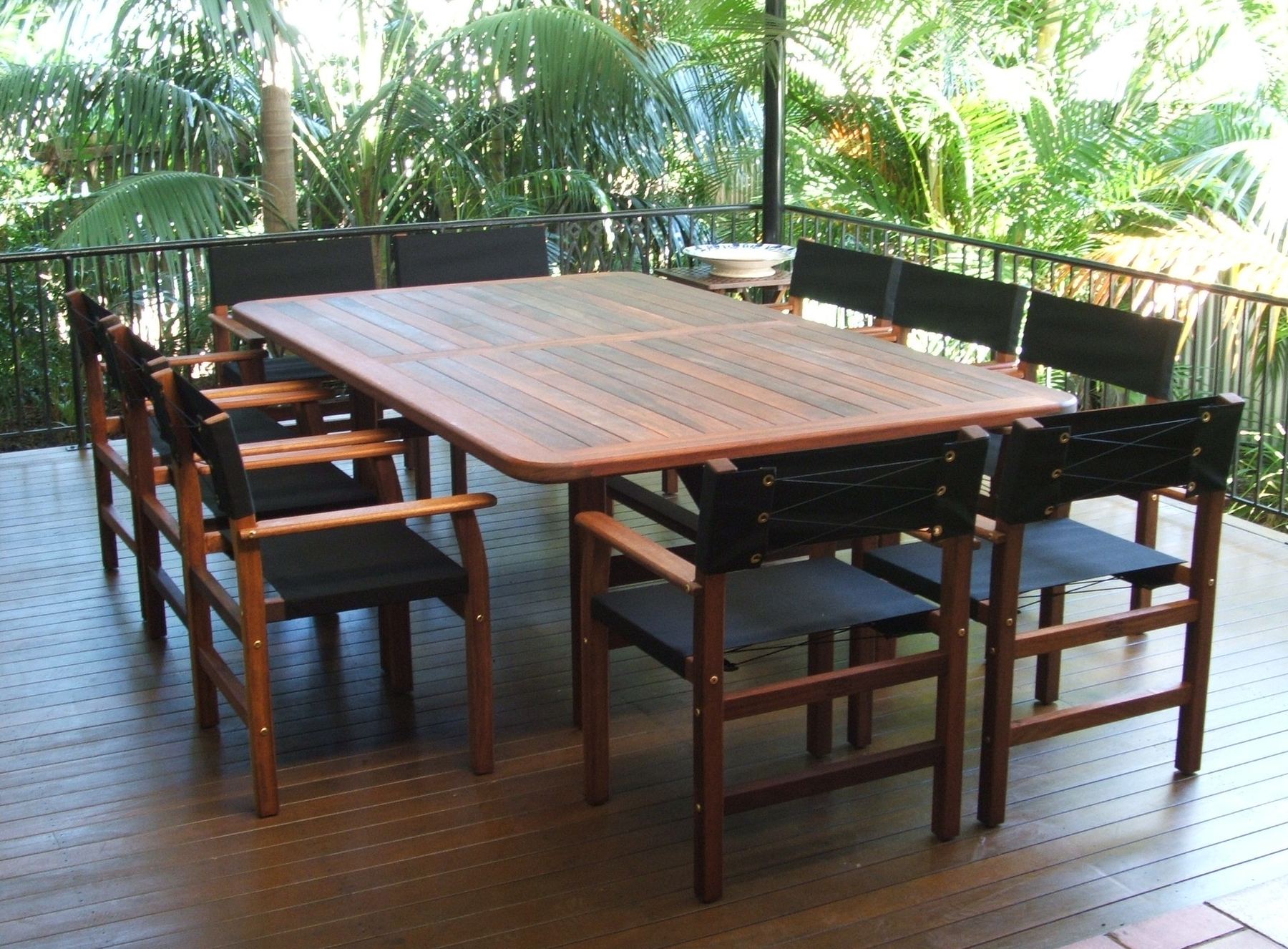 A long Kwila dining table with eight Miami chairs in black canvas on a timber deck under palms